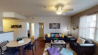 This single-floor residence is thoughtfully laid out across several well-defined spaces, offering a comfortable and functional living environment. The living area serves as the heart of the home, featuring hardwood-style flooring, a ceiling fan with light fixture, and large windows dressed with sheer curtains that allow natural light to fill the room. A decorative geometric wood wall art piece anchors the living space, complemented by modern accent shelving and a warm, eclectic design aesthetic. A dining area sits adjacent, providing a seamless flow for everyday living and entertaining. The galley-style kitchen is efficiently designed with light wood cabinetry, dark granite-style countertops, and white appliances including a range, microwave, dishwasher, and refrigerator. A pantry or utility closet adds additional storage. Both bedrooms are carpeted and each features a ceiling fan, a window with natural views of surrounding greenery, and a mirrored sliding-door closet for ample storage. A dedicated desk area in one bedroom makes it well-suited for study or work. The two full bathrooms are similarly appointed, each offering a single-sink vanity with dark countertops, overhead cabinet storage, a mirror, and a tub-shower combination with tiled surround, providing practical and private spaces for daily routines.