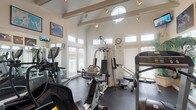 Located at Village Gate in Delaware, Ohio, this well-appointed fitness center offers a bright and energizing environment for a complete workout experience. The room is crowned by a dramatic vaulted ceiling with exposed beams and a circular clerestory window, lending the space an open, airy feel. Track lighting runs along the ceiling, supplementing the generous natural light that pours in through numerous large windows fitted with slatted blinds, offering views of the surrounding exterior while maintaining privacy.

The flooring throughout is a durable dark rubber mat, ideal for high-impact activity and equipment use. Full-length mirrors line multiple walls, visually expanding the space and allowing users to monitor their form during exercise. The cardio section is well-equipped with treadmills — including a Landice model — elliptical trainers, and stationary bikes. A multi-station cable strength machine anchors the strength-training area, complemented by free weights and a weight bench.

Framed sports and nature artwork decorates the upper walls, adding visual interest and energy to the environment. A wall-mounted television and a clock are also present for convenience. Glass-paneled doors connect to an adjacent corridor, providing easy access and maintaining a bright, open atmosphere throughout this thoughtfully designed community fitness facility.