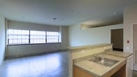 This single-floor space showcases an open-concept layout that seamlessly connects the kitchen and living areas. The living area is anchored by a wide, multi-pane window that draws in natural light, illuminating the polished concrete floors that extend throughout the unit. The clean, neutral-toned walls contribute to a minimalist aesthetic, giving the space a raw, industrial character.

The kitchen is positioned adjacent to the living area and is partially separated by a half-wall with a countertop overhang, which could serve as a breakfast bar or casual dining space. The kitchen features laminate countertops with a neutral stone-like finish, wood-toned cabinetry beneath, and a double-basin stainless steel sink. The layout provides functional workspace while maintaining visual openness to the rest of the unit.

The ceiling is smooth and flat, fitted with a central ventilation vent and recessed lighting points. The overall atmosphere is utilitarian yet spacious, offering a blank canvas well-suited for customization. The concrete floors and industrial detailing lend the space a loft-like quality.