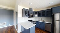 This single-floor space showcases a thoughtfully designed interior with a cohesive color palette and quality finishes throughout. The kitchen serves as a standout feature, outfitted with bold navy blue shaker-style cabinetry — some with glass-front uppers — complemented by striking white quartz countertops with subtle veining. A peninsula provides additional prep space and casual seating potential. Stainless steel appliances, including a gas range, built-in microwave, dishwasher, and refrigerator, complete the well-equipped cooking area. A white mini-tile backsplash adds texture while tying the design together cleanly.

Rich, dark hardwood flooring flows throughout the main living areas, adding warmth and continuity. The living area benefits from vaulted ceilings and a ceiling fan, enhancing the sense of openness. A large window with shutters allows natural light to filter in, while a sliding glass door connects the space to the exterior.

The bathroom is appointed with floor-to-ceiling large-format beige stone-look tile in the walk-in shower, accented by a decorative mosaic border and a coordinating mosaic tile floor, lending a refined, spa-like aesthetic. The muted blue-gray wall tones found throughout the space contribute to a calm, modern ambiance.