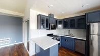 This single-floor space showcases a thoughtfully designed interior with a cohesive color palette and quality finishes throughout. The kitchen serves as a standout feature, outfitted with bold navy blue shaker-style cabinetry — some with glass-front uppers — complemented by striking white quartz countertops with subtle veining. A peninsula provides additional prep space and casual seating potential. Stainless steel appliances, including a gas range, built-in microwave, dishwasher, and refrigerator, complete the well-equipped cooking area. A white mini-tile backsplash adds texture while tying the design together cleanly.

Rich, dark hardwood flooring flows throughout the main living areas, adding warmth and continuity. The living area benefits from vaulted ceilings and a ceiling fan, enhancing the sense of openness. A large window with shutters allows natural light to filter in, while a sliding glass door connects the space to the exterior.

The bathroom is appointed with floor-to-ceiling large-format beige stone-look tile in the walk-in shower, accented by a decorative mosaic border and a coordinating mosaic tile floor, lending a refined, spa-like aesthetic. The muted blue-gray wall tones found throughout the space contribute to a calm, modern ambiance.
