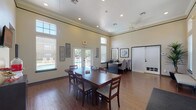 This single-floor community clubroom is designed to serve as a versatile and welcoming gathering space. The interior is defined by notably high ceilings adorned with crown molding and recessed lighting, creating an open and airy atmosphere throughout. Warm hardwood-style flooring extends across the entire space, complementing the neutral, cream-toned walls.

A central dining area anchors the room, furnished with a large dark wood table and cushioned chairs, providing ample seating for group activities or shared meals. Transom windows positioned high along the walls, paired with large ground-level windows fitted with white plantation-style blinds, allow natural light to filter generously into the space. Multiple sets of French doors offer direct access to the outdoors, where views of a pool area are visible, further enhancing the bright and connected feel of the interior.

A dedicated workstation area features a built-in counter with seating and computer access, making the space functional for residents needing a quiet place to work. Additional amenities include a wall-mounted television, a small refreshment station, a ceiling fan, and decorative framed artwork. A secondary door provides access to an adjacent room equipped with a stainless steel refrigerator. The overall design balances comfort and practicality, making this clubroom a well-appointed community amenity.