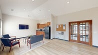 Located at 5617 Muirfield Dr SW in Cedar Rapids, Iowa, this single-floor community space functions as a clubhouse or leasing office, offering a welcoming and functional environment for residents and visitors alike. The open-concept layout is anchored by soaring vaulted ceilings that lend an airy, expansive feel well beyond its physical footprint. Light hardwood-style flooring runs throughout, complementing the neutral, warm-toned walls and dark baseboard trim.

The entry area features large glass double doors that flood the space with natural light, along with warm wood-trimmed windows and a decorative wall clock that add a touch of character to the welcoming foyer. A glass console table and area rug define the entryway transition into the main lounge.

The lounge area is furnished with a sleek sofa, accent chairs, a coffee table, and a wall-mounted flat-screen TV, creating a comfortable gathering space. Multiple windows along the exterior walls provide views of the surrounding property and ample daylight.

A full kitchenette lines one wall, equipped with warm-toned wood cabinetry, granite-style countertops, a sink, a black refrigerator, and a coffee station—ideal for resident amenities. A corkboard information station and French interior doors complete the functional elements of the space, making it both practical and inviting.