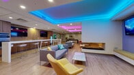 Located at 26600 George Zeiger Drive in Beachwood, Ohio, this expansive single-floor amenity space offers a thoughtfully designed recreational environment spanning a generously sized open floor plan. The space is defined by its striking tiered ceilings outfitted with programmable RGB LED lighting, casting vibrant hues of blue, pink, purple, and orange throughout the interior to create a lively and dynamic atmosphere.

The main lounge area features contemporary modular sofas, accent chairs in bold yellow and neutral tones, a sleek fireplace feature wall, wall-mounted flat-screen televisions, and high-top bar tables with modern stools — ideal for both casual socializing and organized gatherings. An integrated kitchenette area includes warm wood cabinetry, built-in appliances, a large white island with bar seating, and additional TV displays.

The game zone is equipped with a full-size billiards table, a dedicated card and game table surrounded by white chairs, and colorful wall art that adds personality to the space. Light hardwood-style flooring runs throughout, contributing to the open and inviting feel.

A standout feature is the private Full Swing golf simulator room — a darkened, fully enclosed bay complete with a large projection screen, turf hitting area, and a club rack, offering an immersive virtual golf experience.