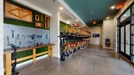 This single-floor bike room is thoughtfully designed to serve cycling enthusiasts with both practicality and style. The centerpiece of the space is a double-tiered bicycle storage system lining one full wall, accommodating a large fleet of branded yellow bikes complete with front baskets. Each bike is neatly organized on heavy-duty vertical racks, maximizing storage capacity without sacrificing accessibility.

Adjacent to the storage racks is a dedicated bike repair and maintenance station, featuring a natural wood workbench and a pegboard wall mounted against a lush artificial green grass backdrop. The pegboard is fully outfitted with a comprehensive array of tools, including wrenches, screwdrivers, and other cycling-specific implements, making on-site maintenance convenient. A floor-standing bike repair stand is also available at the center of the room.

The polished concrete floors run throughout the space, lending an industrial yet clean aesthetic that complements the teal-painted ceiling accents. Large floor-to-ceiling glass garage doors with black steel framing line one side of the room, flooding the interior with natural light and providing open views to the exterior. A wall-mounted digital display screen near the entry offers informational or interactive features for users.

The space also includes a dedicated pet wash or bike wash station finished in gray tile, with a handheld hose and stone flooring for easy cleanup.
