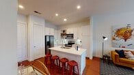 This thoughtfully designed single-floor residence offers a seamless blend of modern functionality and contemporary style. The heart of the home is a well-appointed open-concept kitchen, showcasing crisp white shaker cabinetry extending to the ceiling, a textured tile backsplash, and a full suite of stainless steel appliances including a French door refrigerator, electric range, and built-in microwave. A generous center island with a white countertop and undermount sink provides both prep space and casual seating for several guests. Rich hardwood flooring flows throughout the main living areas, adding warmth and continuity.

The living room is bright and welcoming, bathed in natural light from large windows and French doors that open to an outdoor view. A ceiling fan adds a practical touch overhead, and the open layout allows for flexible furniture arrangements.

Two comfortable bedrooms are finished with plush carpeting and feature windows that invite natural light. One bedroom carries a lively, eclectic character with bold geometric wall art, while the other offers a serene, neutral atmosphere.

The bathroom is clean and modern, featuring a full-length vanity with white cabinetry, a framed mirror, and a soaking tub with shower combination set against dark hardwood flooring, with a walk-in closet conveniently accessible nearby.