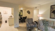 This single-floor residence offers a thoughtfully arranged layout that connects the primary living spaces in a seamless, open-concept flow. The living area is finished with plush carpeting and neutral wall tones, creating a warm and inviting atmosphere. A patterned accent chair and a classic sofa anchor the space, complemented by a glass-top coffee table and a tasteful table lamp that adds a soft, ambient glow. Decorative wall art, including a bold botanical painting and a sculptural wall piece, lend personality and character to the room. The living area opens naturally into the dining space, where a dark-toned dining set provides a defined area for meals and gatherings. A large, striking piece of wall art featuring green pears adds a vibrant pop of color and serves as a focal point in the dining area. Adjacent to the dining space, the kitchen is visible through a connecting passageway, offering convenient access and enhancing the overall open feel of the floor plan. The transition between rooms is marked by tile flooring in the kitchen and entryway areas, contrasting with the soft carpet of the living and dining zones. Overall, the space presents a comfortable, cohesive environment with practical design choices throughout.