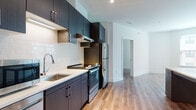 This contemporary single-floor unit showcases a clean, modern design with a cohesive aesthetic carried throughout every room. The kitchen is a standout feature, equipped with dark espresso-toned cabinetry, white quartz countertops, and a classic white subway tile backsplash. Stainless steel appliances — including a refrigerator, electric range, dishwasher, and microwave — complete the well-appointed space, while under-cabinet lighting adds a functional and polished touch. Warm wood-style flooring flows seamlessly from the kitchen into the living areas and bedrooms, creating a sense of continuity and warmth. The bedrooms are bright and airy, benefiting from natural light through large windows, and at least one bedroom includes access to a walk-in closet. The bathroom features a dark wood vanity with a white countertop, an illuminated mirror, and tile flooring in a light stone finish. A separate walk-in glass-enclosed shower adds a spa-like quality to the space. The overall design balances contemporary finishes with a neutral color palette of whites and warm wood tones, resulting in a bright, inviting, and highly functional living environment throughout.