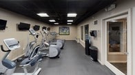 This fitness center offers a comprehensive and well-organized workout environment contained within a single floor. The space features a dark drop-tile ceiling with recessed panel lighting that creates a focused, gym-like atmosphere, while neutral beige walls provide a clean backdrop throughout.

The cardio section is prominently lined with multiple treadmills and elliptical trainers, each positioned for easy access and ample personal space. A row of wall-mounted flat-screen TVs runs along the cardio area, allowing users to stay entertained during their sessions. A large, framed sports action photograph serves as a bold visual centerpiece on the main wall, adding an energetic and motivating character to the space.

The strength and functional training area includes a multi-station cable machine, a recumbent cycle, rowing machines, and a collection of medicine and stability balls in various sizes. Full-length mirrors line one wall, enhancing the sense of space and allowing users to monitor their form during exercise. The flooring throughout is durable rubber gym matting, providing both safety and comfort underfoot.

Additional amenities include a water cooler station, waste receptacles, and a paper towel dispenser, ensuring a clean and convenient experience. A separate doorway leads to an adjoining room, suggesting additional support space. Overall, this fitness center is thoughtfully designed to support a wide range of training activities in a functional and well-maintained environment.