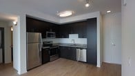 This single-floor residence showcases a clean, modern aesthetic that is immediately apparent in its thoughtfully designed kitchen. The kitchen is appointed with bold, dark flat-front cabinetry that extends floor to ceiling, creating a striking contrast against the light walls and warm hardwood-style flooring. A full suite of stainless steel appliances—including a refrigerator, electric range, built-in microwave, and dishwasher—provides both style and functionality. Granite countertops and a contemporary single-basin sink with a sleek gooseneck faucet complete the workspace, while a simple white tile backsplash keeps the look crisp and uncluttered. The kitchen is laid out in an efficient L-shape, maximizing usable counter and storage space. Overhead, flush-mount and track lighting illuminate the space effectively, contributing to a bright and inviting atmosphere. The warm-toned hardwood-style flooring flows seamlessly throughout, anchoring the contemporary design with an element of understated warmth. Overall, the space presents a cohesive, modern interior with high-quality finishes that balance practicality with refined style.