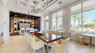 Located at 2130 Leather Fern Dr in Odessa, FL, this single-floor clubhouse facility offers a thoughtfully designed community amenity space combining elegance with functionality. The main lounge area is anchored by a coffered ceiling with a striking sputnik-style chandelier, and features herringbone-patterned light wood flooring that lends warmth to the bright, airy interior. A large communal dining table surrounded by cream upholstered chairs serves as a central gathering point, complemented by smaller café-style tables positioned near floor-to-ceiling windows that flood the space with natural light and provide views of the palm-lined outdoor surroundings. A built-in service counter runs along one wall, equipped with a coffee station, printer, and microwave, while framed line-art pieces add a curated aesthetic touch. Adjoining the lounge is a dedicated private theater room, finished in deep navy blue tones with a dark coffered ceiling and bold geometric-patterned carpet. Rows of white leather recliner seats with cup holders face a large projection screen framed by illuminated built-in shelving displaying decorative accents. A bar-height counter with patterned stools bridges the theater and lounge areas, creating a seamless transition between entertainment and social spaces.