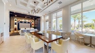 Located at 2130 Leather Fern Dr in Odessa, FL, this single-floor clubhouse facility offers a thoughtfully designed community amenity space combining elegance with functionality. The main lounge area is anchored by a coffered ceiling with a striking sputnik-style chandelier, and features herringbone-patterned light wood flooring that lends warmth to the bright, airy interior. A large communal dining table surrounded by cream upholstered chairs serves as a central gathering point, complemented by smaller café-style tables positioned near floor-to-ceiling windows that flood the space with natural light and provide views of the palm-lined outdoor surroundings. A built-in service counter runs along one wall, equipped with a coffee station, printer, and microwave, while framed line-art pieces add a curated aesthetic touch. Adjoining the lounge is a dedicated private theater room, finished in deep navy blue tones with a dark coffered ceiling and bold geometric-patterned carpet. Rows of white leather recliner seats with cup holders face a large projection screen framed by illuminated built-in shelving displaying decorative accents. A bar-height counter with patterned stools bridges the theater and lounge areas, creating a seamless transition between entertainment and social spaces.