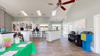 Located at 6 Derby Dr in Hampton, Virginia, this single-floor community space offers a thoughtfully designed open-plan layout that seamlessly integrates a living room, dining room, and kitchen. The interior is defined by dramatic vaulted ceilings that soar overhead, punctuated by skylights that flood the space with natural light. Soft sage-green walls and rich wood-look flooring create a warm, inviting ambiance throughout.

The living area is anchored by a striking floor-to-ceiling herringbone brick fireplace, complemented by large windows that frame views of the surrounding greenery. A ceiling fan with wood-toned blades adds both function and style. The dining area features a substantial wooden table and is flanked by a built-in white bookcase wall unit housing shelving, cabinetry, and a television — a standout architectural feature of the space.

The kitchen runs along one wall with warm wood cabinetry, dark countertops, and a generous window with French door access to an outdoor area. A white wainscoted breakfast bar with bar stool seating serves as a casual dining and gathering spot. The main entry features a full-glass door that welcomes natural light, and elegant French doors with grid panes provide additional interior access, completing this versatile and well-appointed space.