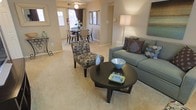 This single-floor residence offers a thoughtfully designed open-concept layout that seamlessly connects the living and dining areas, creating a natural flow ideal for both everyday living and entertaining. The living area is anchored by a plush sofa in soft teal, complemented by a patterned accent chair and a dark round coffee table, all set atop wall-to-wall carpeting in warm beige tones. Neutral walls provide a versatile backdrop that enhances the sense of openness and light throughout the space. A floor lamp adds a warm ambient glow, while decorative accessories — including accent pillows, vases, and tabletop décor — lend the room a polished, curated feel. The adjacent dining area features a dark-finished table with woven chairs and is positioned near a window that invites natural light into the space. A ceiling fan is also present, adding to the comfort and airflow of the home. The entryway, visible from the living area, offers a welcoming transition into the space. The overall ambiance is cozy and inviting, with a cohesive color palette of browns, teals, and warm neutrals tying the living and dining areas together in a harmonious and functional single-floor layout.