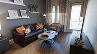 This single-floor space showcases a thoughtfully designed interior that blends contemporary style with warm, inviting character. The living area serves as the focal point, featuring rich, dark hardwood flooring that anchors the room and complements the deep charcoal accent wall. A substantial leather sofa and a coordinating upholstered chair are arranged around a pair of round industrial-style coffee tables, creating a cohesive and functional seating arrangement. Bold orange and chevron-patterned throw pillows inject vibrant pops of color against the neutral tones, while patterned floor-to-ceiling curtains add texture and visual height to the space. A distinctive "TX" wall décor piece and carefully curated shelf art lend the room a strong sense of place and personality. Natural light filters generously through large windows and a glass-paneled door that opens to a balcony, brightening the interior and connecting the space to the outdoors. An industrial-style tripod floor lamp adds both functional lighting and a stylish design element. Overall, the space exudes a modern, urban aesthetic with warm earthy tones, bold accents, and deliberate attention to detail throughout.