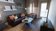 This single-floor space showcases a thoughtfully designed interior that blends contemporary style with warm, inviting character. The living area serves as the focal point, featuring rich, dark hardwood flooring that anchors the room and complements the deep charcoal accent wall. A substantial leather sofa and a coordinating upholstered chair are arranged around a pair of round industrial-style coffee tables, creating a cohesive and functional seating arrangement. Bold orange and chevron-patterned throw pillows inject vibrant pops of color against the neutral tones, while patterned floor-to-ceiling curtains add texture and visual height to the space. A distinctive "TX" wall décor piece and carefully curated shelf art lend the room a strong sense of place and personality. Natural light filters generously through large windows and a glass-paneled door that opens to a balcony, brightening the interior and connecting the space to the outdoors. An industrial-style tripod floor lamp adds both functional lighting and a stylish design element. Overall, the space exudes a modern, urban aesthetic with warm earthy tones, bold accents, and deliberate attention to detail throughout.
