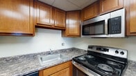 Located at 2100 E High St in Springfield, Ohio, this single-floor residential unit offers a practical and comfortable layout designed for everyday living. The kitchen is well-appointed with warm honey-toned wood cabinetry extending to the upper walls, dark granite-style countertops, stainless steel appliances including an electric range, over-the-range microwave, and dishwasher, along with a stainless steel sink — providing ample workspace and storage. The living area is open and airy, featuring neutral-toned carpeting, wall vents for climate control, and a clear sightline into the kitchen, creating a connected and cohesive feel. The carpeted bedrooms are generously sized, bathed in natural light from large windows that frame outdoor views of the surrounding Springfield area. One bedroom features sliding glass door access to a private balcony with elevated views of the tree-lined surroundings. The bathroom is clean and functional, offering a wide vanity with a marble-style countertop, a large mirror with built-in medicine cabinet storage, a glass-enclosed shower, and a separate water closet. Rich wood-toned interior doors are found throughout, adding warmth to the otherwise light and neutral interior palette.