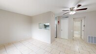 This single-story, 1-bedroom, 1-bathroom unit offers a practical and well-organized layout designed to maximize every square foot of its compact footprint. The living room is the heart of the space, featuring light beige ceramic tile flooring that runs consistently throughout the entire unit, neutral gray walls, and a ceiling fan with an integrated light fixture. A front entry door and a large window dressed with vertical blinds allow natural light to brighten the room. A partial wall with a pass-through opening connects the living area to the kitchen, which features white cabinetry and a mosaic tile backsplash, adding a subtle design accent. The bedroom offers a comfortable and functional retreat, complete with a ceiling fan, a built-in closet with shelving, and a window that brings in additional natural light. The bathroom is finished with matching beige floor tile, a tiled tub and shower surround, and a vanity topped with a marble-patterned countertop. A hallway connects the main rooms, providing a logical flow throughout the unit. The overall aesthetic is clean, neutral, and move-in ready.