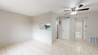 This single-story, 1-bedroom, 1-bathroom unit offers a practical and well-organized layout designed to maximize every square foot of its compact footprint. The living room is the heart of the space, featuring light beige ceramic tile flooring that runs consistently throughout the entire unit, neutral gray walls, and a ceiling fan with an integrated light fixture. A front entry door and a large window dressed with vertical blinds allow natural light to brighten the room. A partial wall with a pass-through opening connects the living area to the kitchen, which features white cabinetry and a mosaic tile backsplash, adding a subtle design accent. The bedroom offers a comfortable and functional retreat, complete with a ceiling fan, a built-in closet with shelving, and a window that brings in additional natural light. The bathroom is finished with matching beige floor tile, a tiled tub and shower surround, and a vanity topped with a marble-patterned countertop. A hallway connects the main rooms, providing a logical flow throughout the unit. The overall aesthetic is clean, neutral, and move-in ready.
