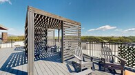 Located at 410 Court Street in Lynchburg, Virginia, this rooftop deck offers an expansive outdoor experience elevated above the city with commanding views of the surrounding treetops, mountains, and skyline. The deck is constructed with durable composite decking in a cool grey finish, providing a clean and low-maintenance surface throughout the space.

A defining architectural feature of the rooftop is the custom wood pergola structure, which incorporates slatted privacy screens on multiple sides, creating semi-enclosed seating alcoves that offer both shade and shelter while maintaining open sightlines to the landscape beyond. Adirondack-style chairs and small accent tables are arranged beneath and around the pergola, providing comfortable gathering areas suited for relaxing or socializing outdoors.

The deck wraps along the building's exterior, which features an impressive floor-to-ceiling glass curtain wall façade with steel framing, allowing natural light to flow between the interior and the outdoor space. Exposed brick accents on the building exterior add an industrial character consistent with the urban setting. Black steel railings line the perimeter of the deck, ensuring safety while preserving the unobstructed panoramic views. Decorative potted plants add touches of greenery throughout. This rooftop deck is a well-appointed outdoor amenity ideal for enjoying Lynchburg's scenic surroundings.