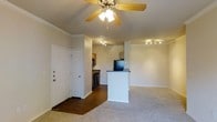 This single-story residence offers a practical and welcoming open-concept floor plan that seamlessly connects the main living area with the kitchen. Upon entry through the solid white front door, you are greeted by a spacious living room finished with plush carpeting and elegant crown molding along the ceiling. A classic ceiling fan with integrated lighting anchors the room, providing both comfort and illumination.

The kitchen is positioned toward the rear of the open space and is defined by a white breakfast bar peninsula with a dark countertop, which serves as both a functional workspace and a casual dining divider. Dark-finish appliances, including a refrigerator and stove, are neatly incorporated into the kitchen layout, which transitions to hardwood-style flooring for easy maintenance. Track lighting on the kitchen ceiling ensures the space is well-lit for cooking and meal preparation.

The neutral color palette throughout — from the beige walls to the light carpeting — creates a clean, cohesive ambiance that is both airy and inviting. The open sightlines between the entry, living area, and kitchen make the space feel connected and functional, ideal for everyday living and entertaining.