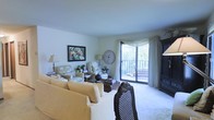 This single-floor residence centers around a comfortable and thoughtfully arranged living room that blends classic charm with functional design. The space is bathed in natural light, courtesy of a large sliding glass door that leads to a private balcony overlooking lush, tree-filled surroundings — a standout feature that brings the outdoors in and adds a sense of openness to the room.

The living room is generously proportioned, accommodating a plush, slipcovered sofa and coordinating seating, including a floral accent chair and an ornate dark-wood dining chair that add visual interest. Soft, neutral-toned carpet grounds the space, complementing the cream and beige color palette throughout. Elegant sheer curtains with a draped valance frame the balcony door, enhancing the room's refined yet relaxed ambiance.

Decorative touches include a large round wall clock, a framed colorful artwork, and warm-toned table lamps that contribute to the inviting atmosphere. A dark wood armoire anchors one corner of the room, providing both storage and a focal point. The overall feel is warm, lived-in, and classic — a comfortable retreat with pleasing natural views.