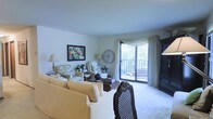 This single-floor residence centers around a comfortable and thoughtfully arranged living room that blends classic charm with functional design. The space is bathed in natural light, courtesy of a large sliding glass door that leads to a private balcony overlooking lush, tree-filled surroundings — a standout feature that brings the outdoors in and adds a sense of openness to the room.

The living room is generously proportioned, accommodating a plush, slipcovered sofa and coordinating seating, including a floral accent chair and an ornate dark-wood dining chair that add visual interest. Soft, neutral-toned carpet grounds the space, complementing the cream and beige color palette throughout. Elegant sheer curtains with a draped valance frame the balcony door, enhancing the room's refined yet relaxed ambiance.

Decorative touches include a large round wall clock, a framed colorful artwork, and warm-toned table lamps that contribute to the inviting atmosphere. A dark wood armoire anchors one corner of the room, providing both storage and a focal point. The overall feel is warm, lived-in, and classic — a comfortable retreat with pleasing natural views.