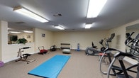 This fitness room offers a comprehensive workout environment designed to accommodate a variety of exercise routines. The space is finished with neutral cream-colored walls and durable carpet flooring, creating a clean and functional atmosphere. Overhead fluorescent lighting provides consistent illumination throughout the room.

The layout is thoughtfully organized to maximize usability. Cardio equipment, including a treadmill and an elliptical trainer, is positioned along one wall, with a large mirror enhancing the sense of space and allowing users to monitor their form. A dedicated open area at the center of the room features a full-length blue exercise mat, ideal for stretching, yoga, or floor-based workouts.

Additional equipment is distributed throughout the room, including a rowing-style machine, an ab roller, a stability ball, a weight bench, a dumbbell rack, and various compact fitness accessories. A cork bulletin board near the entry door provides a space for notices or workout information, and a pass-through window with decorative greenery adds a welcoming touch. The room also includes a separate door for convenient access. Overall, the space is well-organized and equipped to support a broad range of fitness activities.