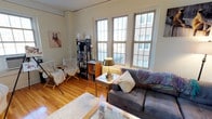 This single-floor urban residence offers a warm and inviting atmosphere, characterized by classic architectural details and thoughtful design. The living area serves as the heart of the space, showcasing rich hardwood flooring that adds warmth and continuity throughout. A striking trio of tall, grid-paned windows floods the room with abundant natural light while framing views of the surrounding cityscape, creating a bright and airy environment. The space features a comfortable seating arrangement, including a plush sofa accented with decorative pillows and a cozy throw, paired with a relaxed folding chair draped with a blanket — ideal for reading or unwinding. A glass-top coffee table and a classic brass table lamp add functional elegance to the room. Built-in and freestanding bookshelves provide ample storage and display space, contributing to the intellectual and lived-in charm of the interior. Soft neutral wall tones complement the natural wood elements, while a plush area rug anchors the seating area. Artwork on the walls adds a personal, curated touch. The overall ambiance is cozy, eclectic, and full of character — a comfortable urban retreat with practical amenities and a welcoming feel.