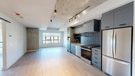 Located in Calgary, AB, this well-designed two-bedroom, two-bathroom unit blends industrial character with modern finishes across a single floor. The open-concept kitchen and living area is anchored by a sleek galley-style kitchen outfitted with slate-grey cabinetry, a tiled backsplash, stainless steel appliances including a gas range, dishwasher, and French door refrigerator, and track lighting beneath an exposed concrete ceiling. Light hardwood flooring runs throughout, lending warmth to the industrial aesthetic. The living area is bright and open, with large grid-pane windows drawing in natural light and a glass door opening to a balcony with city views. A concrete structural column and a sliding barn door add distinctive architectural details to the space. The primary bedroom features a generous walk-in closet with built-in shelving and a large window framing sweeping Calgary skyline views. The ensuite bathroom is finished with dark square wall tiles, a glass-enclosed shower, a wood-toned vanity, and large-format floor tiles. The second bathroom offers a full soaker tub with tiled surround. The unit's thoughtful layout and urban design elements make it a refined and functional city dwelling.