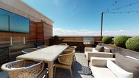 Located at 6851 S Gaylord St in Centennial, Colorado, this exceptional rooftop terrace offers a sophisticated outdoor living experience with sweeping panoramic views of the Rocky Mountains and the surrounding landscape. The expansive terrace is finished with large-format slate-toned porcelain tile pavers that lend a clean, modern aesthetic underfoot.

The space is thoughtfully divided into distinct functional zones. A dedicated outdoor dining area features a glass-top wicker table with woven rattan chairs, ideal for al fresco entertaining. Adjacent to the dining area, a comfortable lounge zone is furnished with contemporary upholstered outdoor sofas and chairs, creating an inviting setting for relaxation.

The outdoor kitchen station serves as a standout feature, incorporating a built-in stainless steel grill, a prep counter with stone veneer detailing, and a wall-mounted flat-screen TV — all framed by warm horizontal hardwood slatted privacy screens that add architectural texture and visual warmth. Manicured boxwood topiaries and raised stone planters introduce lush greenery, softening the modern hardscape.

String lights are strung overhead, adding an inviting ambiance for evening gatherings. Access to the terrace is provided through a sleek glass-and-steel door. The open sky exposure and unobstructed mountain vistas make this rooftop an outstanding feature of the property.