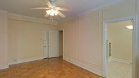 This single-floor space exudes a classic charm through its thoughtfully preserved architectural details. The main living area immediately draws attention with its warm parquet hardwood flooring, arranged in a traditional basket-weave pattern that adds visual texture and character underfoot. Crown molding lines the perimeter of the ceiling, lending an elegant finishing touch to the room's upper edges.

The walls feature decorative raised panel molding, providing subtle architectural depth and a refined aesthetic throughout the space. A ceiling fan with an integrated light fixture is centrally positioned, offering both ambient lighting and airflow for year-round comfort.

The room benefits from a clean, open layout that allows for versatile furniture arrangements. Doorways lead to additional areas of the space, including what appears to be a closet with double doors, providing practical storage solutions. Wide baseboards complement the overall traditional design language present throughout.

The neutral, light toned walls create a bright and airy atmosphere, enhancing the sense of openness. The combination of classic flooring, paneled walls, and detailed moldings gives this space a timeless quality that balances both comfort and style in a cohesive, well-maintained interior.