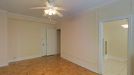 This single-floor space exudes a classic charm through its thoughtfully preserved architectural details. The main living area immediately draws attention with its warm parquet hardwood flooring, arranged in a traditional basket-weave pattern that adds visual texture and character underfoot. Crown molding lines the perimeter of the ceiling, lending an elegant finishing touch to the room's upper edges.

The walls feature decorative raised panel molding, providing subtle architectural depth and a refined aesthetic throughout the space. A ceiling fan with an integrated light fixture is centrally positioned, offering both ambient lighting and airflow for year-round comfort.

The room benefits from a clean, open layout that allows for versatile furniture arrangements. Doorways lead to additional areas of the space, including what appears to be a closet with double doors, providing practical storage solutions. Wide baseboards complement the overall traditional design language present throughout.

The neutral, light toned walls create a bright and airy atmosphere, enhancing the sense of openness. The combination of classic flooring, paneled walls, and detailed moldings gives this space a timeless quality that balances both comfort and style in a cohesive, well-maintained interior.
