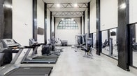 This contemporary fitness center showcases a striking industrial aesthetic, combining clean white walls with bold black steel columns and exposed overhead trusses that draw the eye upward to the soaring ceiling. A dramatic arched window at the far end of the space floods the room with natural light and offers a glimpse of lush greenery beyond, creating an energizing atmosphere for workouts. The polished concrete flooring complements the space's modern-industrial design while remaining highly functional and durable. The facility is well-equipped with a comprehensive lineup of cardio machines, including treadmills and ellipticals, neatly arranged along one side. Strength training is well-represented with cable machines, a dedicated free weight rack with dumbbells, and various resistance stations. Wall-mounted mirrors enhance the sense of openness and allow users to monitor their form, while a flat-screen television adds convenience and entertainment. Pendant and recessed lighting supplements the natural light, ensuring the space remains bright and well-illuminated at all times. The overall layout is spacious and thoughtfully organized, providing ample room for movement between stations and creating an inviting environment for a complete workout experience.