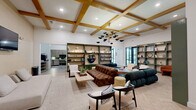 Located at 6490 Iron Horse Blvd in North Richland Hills, TX, this lounge and kitchen space offers a well-appointed, single-floor layout designed for comfort and social interaction. The living area makes an immediate impression with its striking coffered ceiling accented by warm wood beams and a bold mid-century modern chandelier featuring a multi-arm brass and black fixture. Herringbone-patterned hardwood flooring flows throughout, grounding the space with texture and warmth. Built-in shelving units in a sage green finish line the walls, providing ample display space for books and décor. Seating is plentiful and varied, with a tufted brown leather sofa, deep teal sectional, and an assortment of accent chairs arranged around a sculptural wooden coffee table. The entry features a bold navy double door, exposed wood beam ceiling, and a welcoming sitting area. The kitchen is equally impressive, boasting soaring ceilings with exposed wood beams, a generous center island with white quartz countertops, and extensive sage green cabinetry. Stainless steel appliances, a green subway tile backsplash, and globe pendant lighting complete the culinary space. Large barn-style sliding doors and glass exterior doors allow natural light to pour in, enhancing the open, airy ambiance throughout.