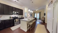 Located at 1201 Central Haven Dr in Mount Pleasant, South Carolina, this well-appointed single-floor residence combines modern finishes with a comfortable, functional layout. The open-concept kitchen serves as the heart of the home, showcasing an expansive granite-topped island with seating for multiple guests, rich dark espresso cabinetry, stainless steel appliances, and elegant pendant lighting. Track lighting and crown molding throughout add a refined architectural touch, while durable hardwood-style flooring flows seamlessly across the main living areas. The living room offers a cozy, well-lit retreat with natural light streaming through large windows dressed with stylish drapes. The bedrooms are carpeted and thoughtfully appointed, each featuring ceiling fans and generous windows. The primary bathroom includes a dual-sink granite vanity with dark cabinetry and direct access to a walk-in closet with built-in shelving. Secondary bathrooms mirror the same high-quality finishes, including granite countertops, dark vanities, and tub-shower combinations. A dedicated in-unit laundry area with side-by-side washer and dryer adds everyday convenience. The overall aesthetic is cohesive and modern, with warm neutral tones creating an inviting atmosphere throughout.