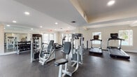 This fitness center is a comprehensively outfitted single-floor facility designed to support a wide range of workout routines. The space is organized into distinct functional zones, each dedicated to a specific type of training. The strength training area houses a Life Fitness Smith machine with an adjustable bench and an assortment of weight plates, alongside multiple selectorized cable machines suited for targeted muscle training. A dedicated free weights section features a full rack of rubber hex dumbbells in graduated sizes, accompanied by a utility bench and a wall-length mirror etched with motivational text. The cardio zone is well-stocked with Life Fitness treadmills, elliptical trainers, and upright bikes, positioned near glass doors and windows that invite ample natural light and outdoor views. Rubber flooring spans the entire space, providing durability and impact absorption throughout. Recessed lighting, crown molding, and neutral-toned walls contribute to a clean, polished aesthetic. Multiple sets of glass-paned doors offer convenient access points and enhance the open, airy feel of the facility. The layout allows for generous spacing between equipment, ensuring comfortable and safe use throughout the space.