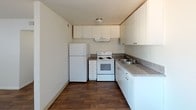 This single-floor space presents a clean and functional layout designed for everyday living. The kitchen serves as a central feature, showcasing white shaker-style cabinetry that extends to the ceiling, maximizing storage capacity. Granite-look laminate countertops line the walls in an efficient L-shaped configuration, providing ample workspace for meal preparation. White appliances — including a refrigerator, electric coil range with oven, and an under-cabinet range hood — complement the bright, cohesive aesthetic of the kitchen. A double-basin stainless steel sink with a standard faucet is conveniently positioned along the counter. The kitchen flooring consists of warm, dark wood-look planks that contrast nicely with the white cabinetry, adding a touch of warmth to the space. Ceiling-mounted flush lighting provides even illumination throughout. The overall design prioritizes practicality and efficiency, with a neutral color palette that creates a light and airy atmosphere. The thoughtful use of vertical cabinetry and counter space makes this kitchen well-suited for comfortable, day-to-day use.