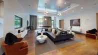 This well-appointed single-floor lounge space combines modern design with functional versatility. The centerpiece of the space is a generously sized open living area anchored by a large dark leather sectional sofa, accented with decorative pillows and set atop a neutral area rug. Overhead, a tray ceiling with recessed lighting and a pair of striking sputnik-style gold chandeliers adds a sophisticated, mid-century modern flair to the room. Warm hardwood flooring flows throughout, complementing the clean white walls and contemporary furnishings.

Floor-to-ceiling windows with patterned drapery allow ample natural light to filter in, offering views of lush greenery outside. A built-in media console and wall-mounted artwork add refined touches to the lounge area.

Adjacent to the main seating space is a bar-height counter with seating, ideal for casual gathering or work. A dedicated kitchenette nearby features dark cabinetry, subway tile backsplash, a stainless steel sink, and under-counter appliances, providing convenient service capabilities for the space.

A standout architectural feature is a glass-walled private room with wood-framed frosted glass panels, accessible via a full-height sliding glass door. This enclosed area offers a quieter, semi-transparent retreat within the broader open-plan layout, making the space well-suited for both social and focused use.