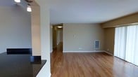 This single-floor space offers an open and functional layout with warm hardwood flooring throughout, complemented by neutral beige-toned walls and white trim that create a clean, understated aesthetic. The main living area is generously proportioned, providing ample room for furniture arrangement, and is served by a wall-mounted HVAC vent, ensuring year-round comfort. Recessed architectural details and structural columns subtly define the transition between the living area and the adjacent kitchen space, which features dark granite countertops visible along the edge of the frame, offering a striking contrast to the lighter surroundings. A hallway extends from the main living space, illuminated by a ceiling-mounted light fixture, and leads to additional rooms beyond, suggesting a practical and well-organized floor plan. The textured white ceilings add a classic finish to the interior, while the consistent flooring unifies the space visually. Overall, the space presents a straightforward, move-in-ready environment with a neutral palette that provides flexibility for a variety of interior design styles.