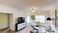 This single-floor space offers a thoughtfully designed living environment that balances comfort and functionality. The living area serves as the heart of the home, featuring plush wall-to-wall carpeting and a ceiling fan with integrated lighting that keeps the room comfortable year-round. Large windows fitted with blinds flood the space with abundant natural light, creating a bright and airy atmosphere throughout the day. A sliding glass door provides additional light and easy access to the outdoors. The room is well-suited for relaxation and entertainment, with ample space to accommodate a large flat-screen television set atop a stylish media console with decorative lattice-front cabinets for storage. Seating arrangements include a curved accent chair with a matching ottoman, offering a cozy reading or lounging nook. A striped area rug in warm tones anchors the space and adds a layer of visual interest to the carpeted floor. Neutral wall tones and warm artificial lighting complement the natural light, contributing to an overall inviting and serene ambiance that makes this space a comfortable and practical place to call home.
