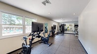 This fitness center offers a thoughtfully arranged workout space across a single floor, providing ample room for a comprehensive range of exercise activities. The facility features durable rubber tile flooring throughout, designed to withstand heavy equipment and high-traffic use while providing cushioning underfoot.

The cardio section is well-appointed with multiple Echelon smart stationary bikes equipped with large interactive screens, a treadmill, and an elliptical trainer. Full-length mirrors line one wall, enhancing the sense of space and allowing users to monitor their form during workouts. A multi-station cable machine and a dedicated free weights area with a TRX-branded dumbbell rack — spanning a range of weights — round out the strength training options. A flat weight bench accompanies the free weights section.

Natural light fills the space through multiple windows, several of which overlook an outdoor pool area with lounge seating and shaded umbrellas, creating an energizing atmosphere. Recessed ceiling lighting provides consistent illumination throughout. A wall-mounted television is present near the entry area, and an additional monitor is installed near the free weights section. The facility includes clearly marked exits and multiple entry points, including a set of French doors, ensuring ease of access.