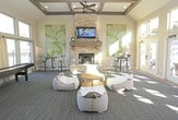 Located at 6610 34th St in Lubbock, Texas, this single-story community clubhouse is designed to serve as a welcoming social hub for residents. The main lounge area is defined by soaring vaulted ceilings accented with exposed wooden beams, creating an open and airy atmosphere. A floor-to-ceiling stacked stone fireplace with a wood mantel serves as the room's focal centerpiece, complemented by a wall-mounted television above it — ideal for gatherings and relaxation.

The lounge is generously lit by an expansive wall of French doors topped with transom windows, offering sweeping views of the surrounding outdoor courtyard and community grounds. Natural light floods the interior throughout the day. Comfortable seating, including bean bag chairs and a round accent table, provides a casual, flexible layout. A shuffleboard table and bar-height bistro seating add recreational and social appeal.

An adjacent area features glass-paned interior French doors that connect to a kitchenette or refreshment space, visible with a stainless steel refrigerator and additional counter space. The entry area transitions smoothly between wood-look tile flooring and plush carpeting. A workspace with a desk and computer provides a functional area for residents. Throughout, the design balances contemporary finishes with warm, inviting details suited for community use.