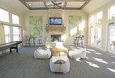Located at 6610 34th St in Lubbock, Texas, this single-story community clubhouse is designed to serve as a welcoming social hub for residents. The main lounge area is defined by soaring vaulted ceilings accented with exposed wooden beams, creating an open and airy atmosphere. A floor-to-ceiling stacked stone fireplace with a wood mantel serves as the room's focal centerpiece, complemented by a wall-mounted television above it — ideal for gatherings and relaxation.

The lounge is generously lit by an expansive wall of French doors topped with transom windows, offering sweeping views of the surrounding outdoor courtyard and community grounds. Natural light floods the interior throughout the day. Comfortable seating, including bean bag chairs and a round accent table, provides a casual, flexible layout. A shuffleboard table and bar-height bistro seating add recreational and social appeal.

An adjacent area features glass-paned interior French doors that connect to a kitchenette or refreshment space, visible with a stainless steel refrigerator and additional counter space. The entry area transitions smoothly between wood-look tile flooring and plush carpeting. A workspace with a desk and computer provides a functional area for residents. Throughout, the design balances contemporary finishes with warm, inviting details suited for community use.