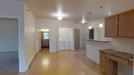 This single-floor space is designed with an open-concept layout that seamlessly connects the main living area to the kitchen. The living area features light-toned hardwood-style flooring that runs throughout, contributing to a bright and airy atmosphere. Overhead, a flush-mount ceiling light provides ambient illumination, while the smooth, neutral-toned walls offer a clean, versatile backdrop.

The kitchen is positioned to one side and is defined by a granite-topped peninsula with cabinetry below, providing both prep space and a casual dining or entertaining surface. Two pendant lights hang above the peninsula, adding focused task lighting. The countertop continues along the kitchen wall, offering ample workspace.

A dedicated laundry closet is tucked conveniently within the unit, housing a full-sized side-by-side washer and dryer. Adjacent to the main space, a bathroom is visible, featuring tile flooring, a vanity with cabinetry, and a window that introduces natural light. The overall layout is practical and efficient, making thoughtful use of the available space while maintaining a cohesive and livable interior environment.