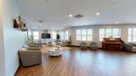 Located at 20 Day St in Norwalk, Connecticut, this single-floor community space offers a well-appointed and versatile layout designed for both relaxation and social interaction. Warm hardwood-style flooring runs throughout, unifying the open floor plan and contributing to a welcoming, contemporary ambiance.

The living area is generously proportioned, featuring soft light-blue accent walls, recessed lighting, and multiple double-hung windows that flood the space with natural light. Comfortable seating arrangements, including plush sofas and rounded accent chairs, are thoughtfully arranged around a central coffee table, creating an inviting lounge environment. A flat-screen television anchors one side of the room, while decorative wall shelving adds a personalized touch.

The kitchen and dining area is equally impressive, showcasing crisp white cabinetry with teal tile backsplash accents, stainless steel appliances including a full-size refrigerator, and a large central dining table surrounded by modern chairs. A striped area rug defines the dining zone, adding a pop of color and texture.

A separate open area provides additional flexible space, currently featuring a shuffleboard table ideal for recreational use. The space also includes a utility corridor with access doors, ensuring smooth circulation throughout. Overall, this thoughtfully designed space balances functionality with a clean, modern aesthetic.