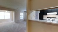 This single-floor residence offers a practical and comfortable layout designed for everyday living. The living area is finished with soft wall-to-wall carpeting and painted in warm neutral tones, creating a welcoming and versatile environment. A window at the far end of the living space allows natural light to filter in, brightening the room and offering a view of the exterior. The textured ceiling adds a subtle architectural detail throughout the space. A defining feature of the layout is the pass-through window connecting the kitchen to the living area, complete with a white countertop ledge that functions as a convenient serving or breakfast bar. The kitchen is equipped with dark-finish appliances, including a full-size refrigerator, providing a striking contrast to the lighter walls and cabinetry. This pass-through design enhances the sense of openness and connectivity between the kitchen and main living space. A hallway visible in the background suggests additional rooms beyond, contributing to the overall functional flow of the floor plan. The neutral color palette and open arrangement make the space adaptable to a variety of furnishing styles and personal tastes.