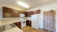 This single-floor unit offers a functional and comfortable layout designed for everyday living. The kitchen is a standout feature, showcasing rich, dark-stained wood cabinetry that extends to the upper walls, providing generous storage throughout. The L-shaped counter configuration offers ample workspace, complemented by light-toned countertops, a double-basin stainless steel sink, and a full suite of white appliances including a refrigerator, electric range, over-the-range microwave, and dishwasher. Durable hardwood-style flooring runs through the kitchen and living areas, adding warmth and visual continuity. The living area is bright and open, featuring a sliding glass door that leads to a private balcony with outdoor views, allowing natural light to fill the space. A wall-mounted air conditioning unit and baseboard heating provide year-round climate control. The bedroom is finished with plush carpet flooring and includes a window that frames views of the surrounding neighborhood, with baseboard heating ensuring comfort. The bathroom is clean and straightforward, featuring a full soaking tub with shower, a curved curtain rod, and crisp white finishes throughout. The entry door features a classic six-panel wood design, creating a welcoming arrival point to the unit.