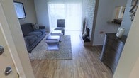 This single-floor space is defined by its warm, inviting character and thoughtful design details. The living area serves as the centerpiece, showcasing rich hardwood-style flooring that extends throughout, lending a cohesive and polished look to the space. A striking white brick fireplace anchors one side of the room, providing a classic architectural focal point that adds both charm and texture to the interior.

Large windows dressed with vertical blinds allow an abundance of natural light to pour in, creating a bright and airy atmosphere. The living area is generously proportioned, accommodating a comfortable seating arrangement that includes a plush sofa, accent chairs, and a set of coordinating ottomans arranged over a shag area rug — all contributing to a relaxed yet stylish ambiance.

The neutral color palette of grays and warm wood tones ties the space together, offering a versatile backdrop suitable for a variety of décor styles. The open layout promotes a seamless flow between areas, enhancing the sense of spaciousness throughout. Overall, this residence combines functional living with timeless design elements, making it a comfortable and welcoming environment.