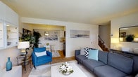 This two-story residence offers a thoughtfully designed layout that balances comfort and functionality across both levels. The main floor centers around an inviting living area, where light-toned walls and neutral flooring create a clean, airy atmosphere. A stylish seating arrangement anchors the space, complemented by decorative wall art that adds a contemporary touch. A staircase with a classic railing leads to the upper level, providing a natural focal point within the room.

The living area flows seamlessly into an open-concept dining and kitchen space, connected through a wide pass-through opening that enhances the sense of space. The kitchen features white cabinetry, a pendant light over the dining area, and warm wood-toned flooring that distinguishes it from the main living space. The dining area accommodates a compact table with seating, making it ideal for everyday meals.

Throughout the main level, an abundance of natural light filters in, brightening the interiors and highlighting the clean lines of the design. Lush indoor plants add a refreshing, natural element to the décor. Overall, this home presents a bright, well-connected layout with a welcoming ambiance suitable for comfortable everyday living.